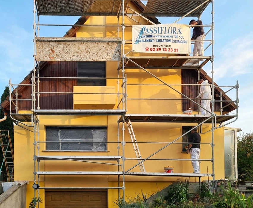 Chantier de ravalement de façade en extérieur, deux ouvriers au travail : l'un accroupi au sol, l'autre sur échafaudage tubulaire