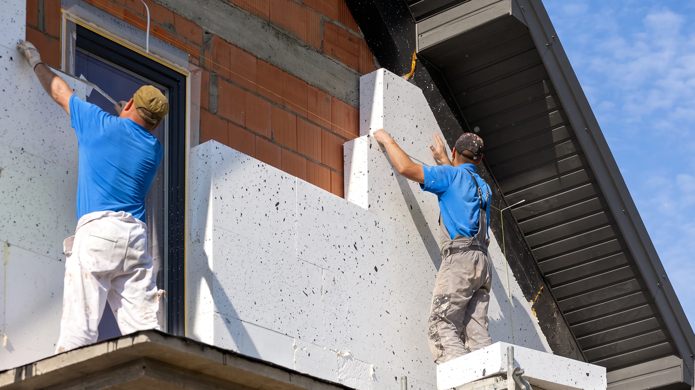 Deux ouvriers en gilets jaunes haute visibilité installent des panneaux d'isolation thermique extérieure sur façade de maison individuelle